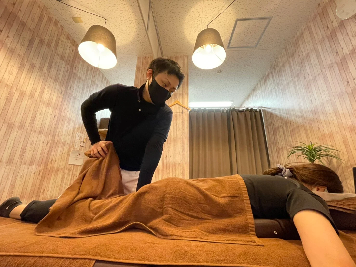 木目調の壁に囲まれた施術室で、黒いマスクの施術者がうつ伏せの女性にかけたタオルを整えながら施術する様子